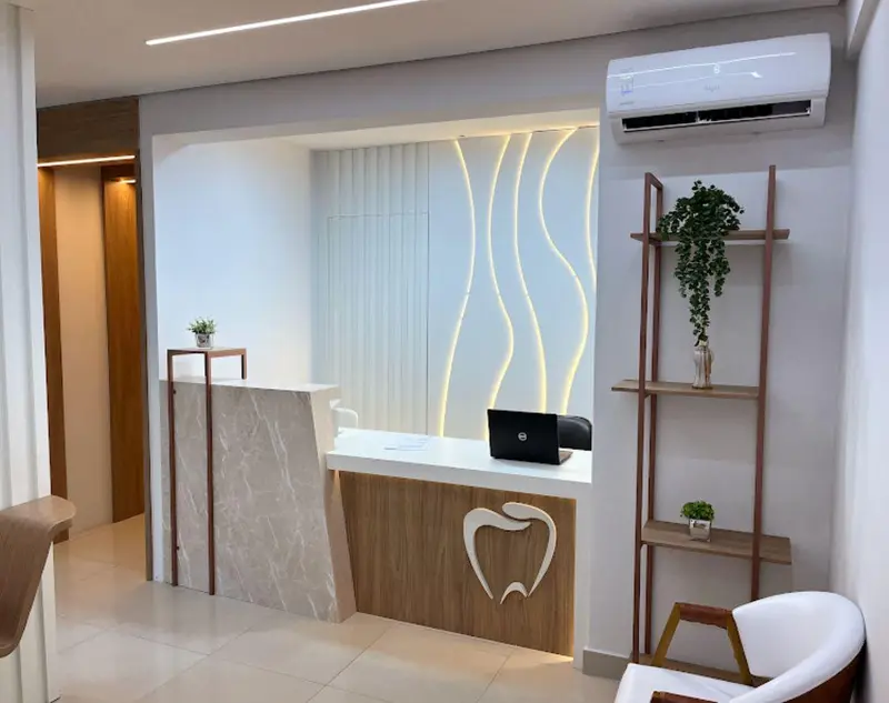 Recepção com balcão de mármore e madeira, parede com ondas de luz embutida, estante com plantas e poltrona branca